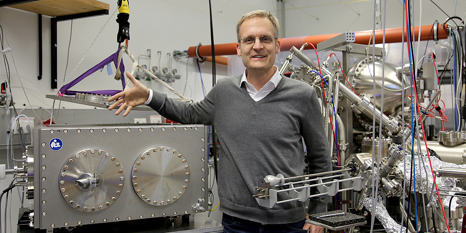 A man with glasses and short blond hair stands in the midst of shiny silver equipment. He points to a metal square box on which two large circles can be seen.