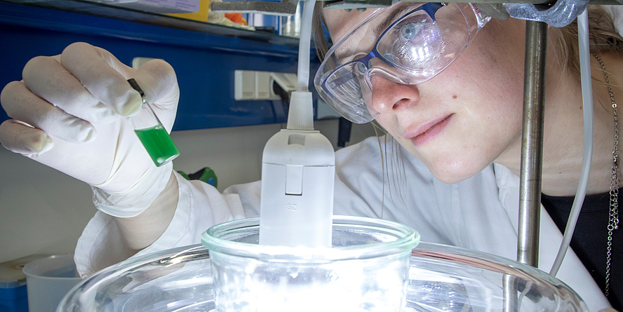 Close-up of a woman wearing laboratory glasses looking at a small bottle of green liquid over a light source.