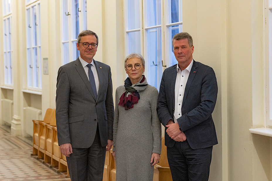 Drei Personen blicken in die Kamera, links und rechts stehen zwei Männer, in der Mitte eine Frau. Sie sind formal gekleidet.