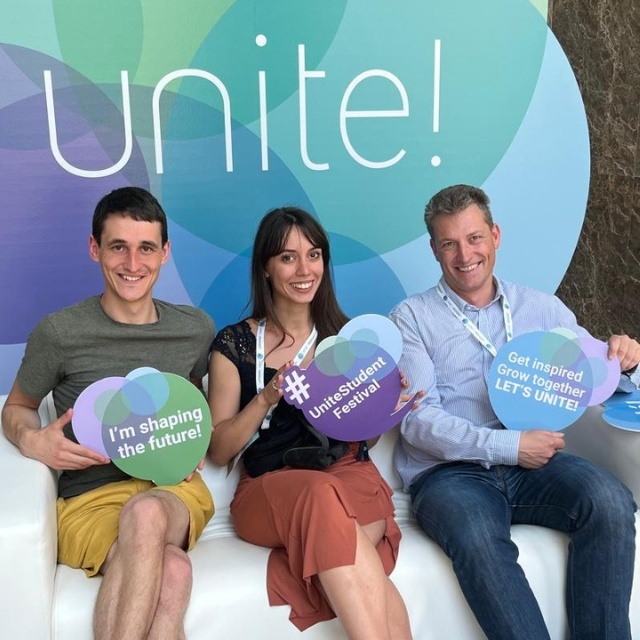 Drei Studierende halten bunte Schilder der Universitätenallianz „Unite!“ hoch.