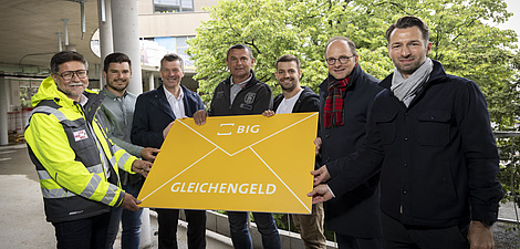 Four men are standing on a building site holding a large envelope in their hands.