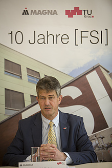 Mann mit dunklen Haaren, schwarzen Anzug und gelber Krawatte sitzt vor einem Plakat, darauf steht 10 Jahre FSI.