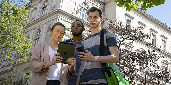 Eine junge Frau und zwei junge Männer stehen vor einem Gebäude. Sie halten ein Tablet und ein Smartphone in der Hand. Bildquelle: Lunghammer – TU Graz
