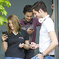 Zwei junge Männer und eine junge Frau stehen lachend beieinander und schauen auf ihre Mobiltelefone.