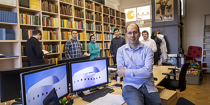 Michael Kerber sitzt auf einem Schreibtisch in einer kleinen Bibliothek. Hinter ihm steht seine Arbeitsgruppe.