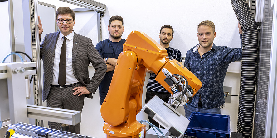 Several men stand behind an orange robot arm.