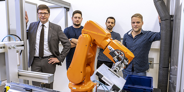 Mehrere Männer stehen hinter einem orangen Roboterarm.