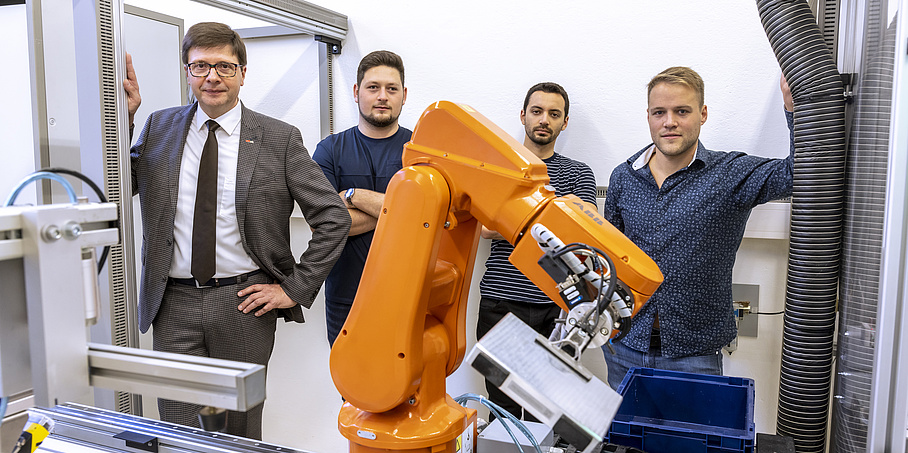 Mehrere Männer stehen hinter einem orangen Roboterarm.