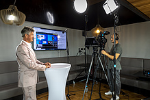 Aufnahmesituation in einem Studio: Rechts Kameramann mit Kamera, links Mann mit hellem Anzug, der gerade in die Kamera spricht. 