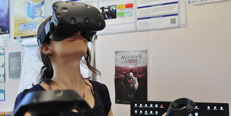 Eine junge Frau hat eine große schwarze Virtual-Reality-Brille aufgesetzt und hält in jeder Hand einen schwarzen Joystick