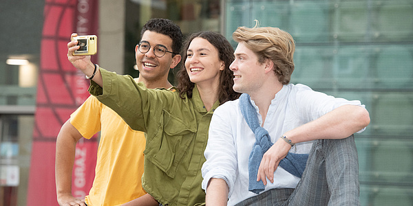 Two men and a woman are sitting close together, taking a selfie with their mobile phone.