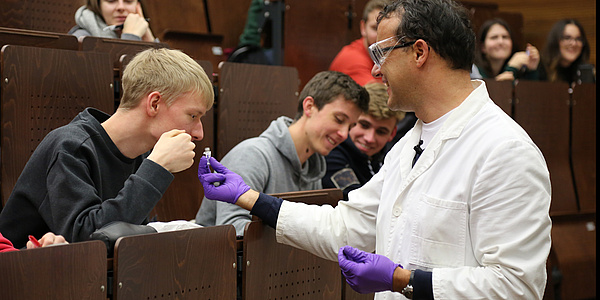 A professor wearing safety glasses and protective gloves shows a laboratory sample to students. Source: Baustädter – TU Graz