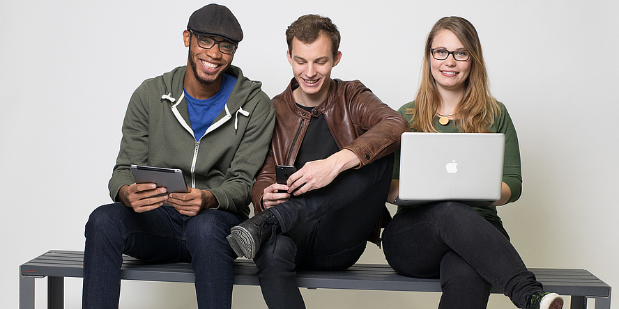 Zwei Burschen udn ein Mädchen sitzen auf einer Bank. Das Mädchen ganz rechts hat einen Laptop am Schoß.