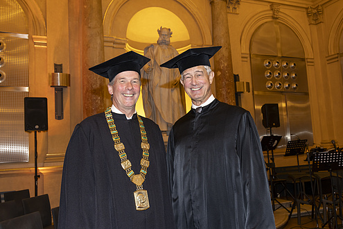 Two men dressed in robes stand next to each other and look into the camera.