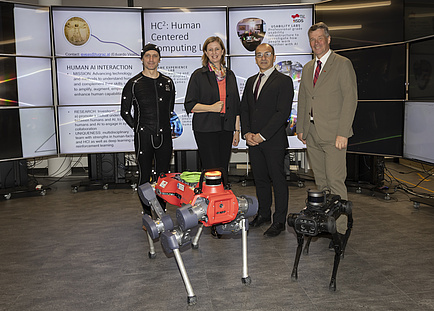 Four people stand behind two dog-like robots and look into the camera