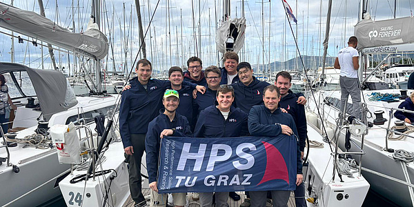 Zehn junge Männer stehen auf einem Segelboot, das im Hafen angelegt hat, und halten ein Transparent mit der Aufschrift "HPS TU Graz".