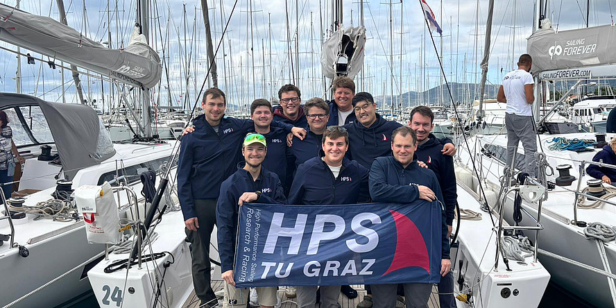 Zehn junge Männer stehen auf einem Segelboot, das im Hafen angelegt hat, und halten ein Transparent mit der Aufschrift "HPS TU Graz".