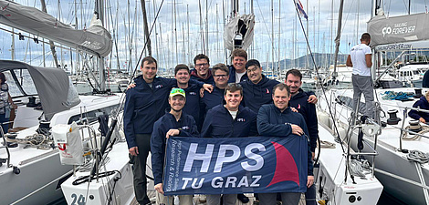 Ten young men stand on a sailing boat moored in the harbour, holding a banner that reads HPS TU Graz.