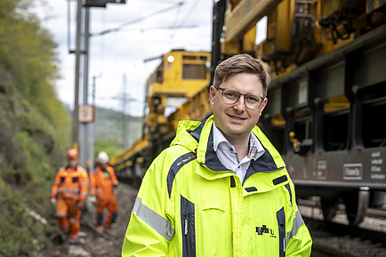 Ein lächelnder Mann, im Hintergrund ein Fahrzeug auf Schienen und Männer in Sicherheitskleidung.