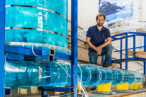 Ein junger Mann mit Bart in Jeans und blauem T-Shirt sitzt im Wasserbaulabor und ist umgeben von Versuchanordnungen in Form von wassergefüllten Plexiglas-Röhren