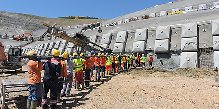 Großbaustelle bei einer betonierten Böschung, vor der viele Arbeiter und Großgeräte zu sehen sind