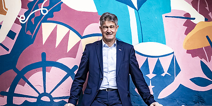 Mann mit blauen Anzug sitzt vor einer bunt bemalten Wand 