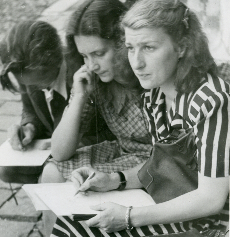 A young man with suit and shirt with pencil, leaning over a sheet of paper, and two young women in short-sleeved dresses; the one on the right holds papers and a pencil and is looking straight ahead. All three are sitting on chairs outside.