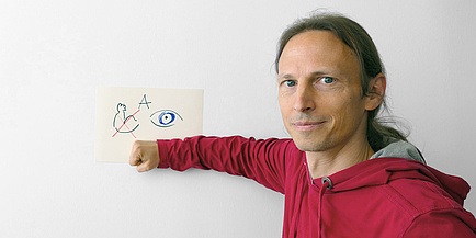 Ein Mann drückt mit seinem Arm einen Zettel mit einem A, einem gebeugten Arm und einem Auge darauf auf eine Tafel.