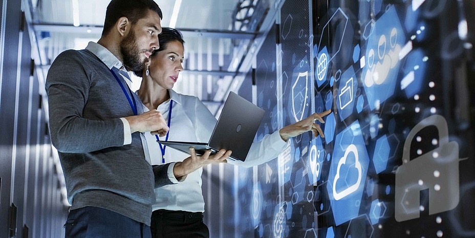 Ein Mann und eine Frau zeigen auf eine digitale Wand, der Mann hat ein Notebook in der Hand
