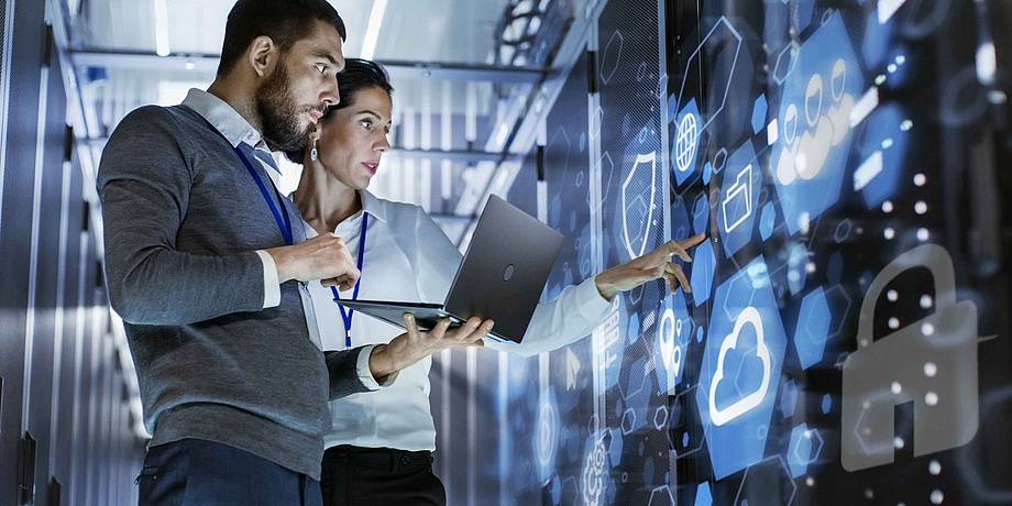 Ein Mann und eine Frau zeigen auf eine digitale Wand, der Mann hat ein Notebook in der Hand