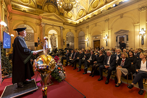 A man stands at the lectern in a festively decorated room and speaks to the audience.