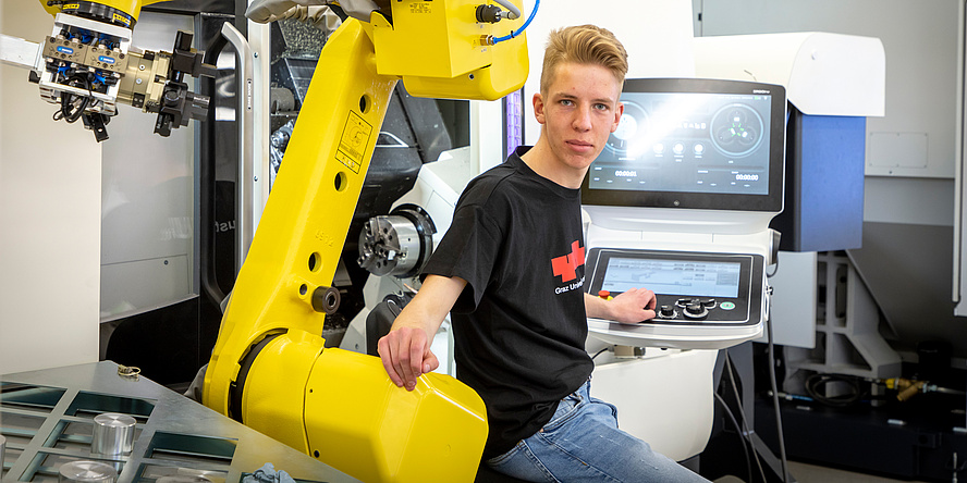 Junger, blonder Mann mit schwarzem Shirt bedient linkerhand einen Computer mit Screen und legt die rechte Hand auf den gelben riesigen Arm einer Maschine.