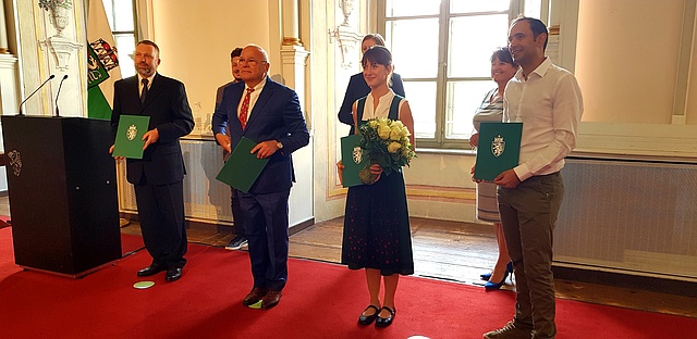 In einem Festsaal stehen die 4 Preisträger in der 1. Reihe und halten eine Urkunde (grün mit steirischem Wappen) in der Hand
