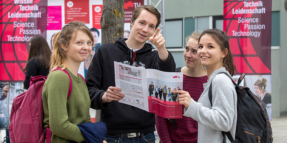 Ein Student erklärt drei Schülerinnen etwas anhand einer Infobroschüre und richtet den Zeigefinger in Richtung Betrachterin oder Betrachter. Im Hintergrund rote Fahnen mit der Aufschrift „Wissen, Technik, Leidenschaft“.