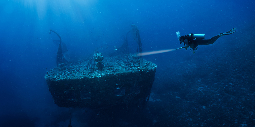A diver shines a torch on a coral-covered wreck.