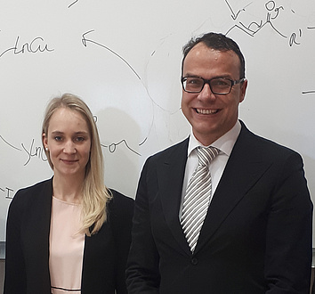 A young woman with blond long hair and a man in a suit and tie are standing next to each other in front of a whiteboard with chemical formulas.