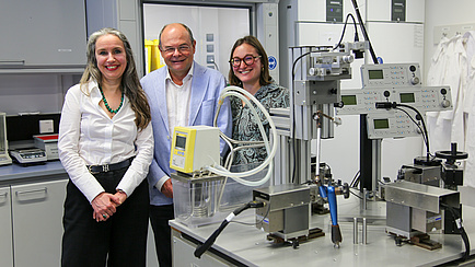 Three people, two women and a man, are standing in a laboratory and smile at the camera. The man is standing in the middle.