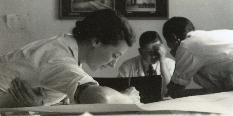 Seitenansicht von Anna Simidoff schreibend oder zeichnend über einen Tisch mit Blättern gebeugt, im Hintergrund zwei junge Männer zueinander gebeugt, ebenfalls konzentriert arbeitend.