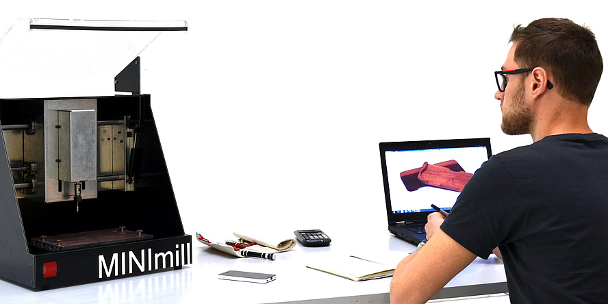 Der Student Jakob Neuhauser sitzt am Schreibtisch vor seinem Laptop, links am Tisch befindet sich die MINImill Desktop-Fräsmaschine.
