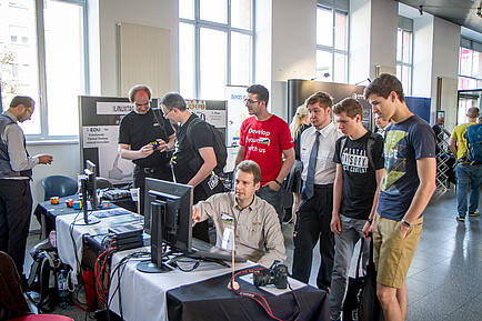 Information booths during the Linux Days. A man is demonstrating something on a computer and is being watched by visitors standing around.