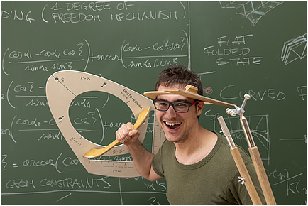 Ein lachender Mann im grünen T-Shirt steht vor einer grünen Tafel, auf der mathemaitsche Formeln geschrieben stehen und hält einen überdimensionalen Zirkel und eine Schablone für Kegelschnitte in den Händen.