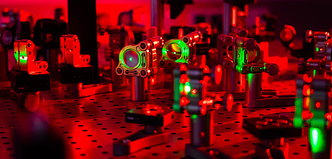 An arrangement of lenses on a laboratory bench illuminated by red light. Green laser light passes through some of the lenses.