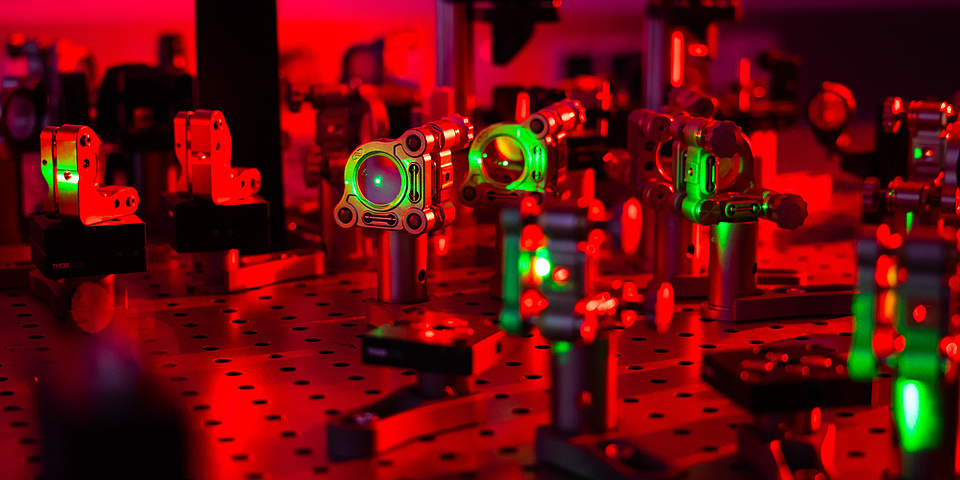 Aufbau von Linsen auf einem Labortisch, der mit rotem Licht ausgeleuchtet ist. Grünes Laserlicht geht durch einige Linsen.