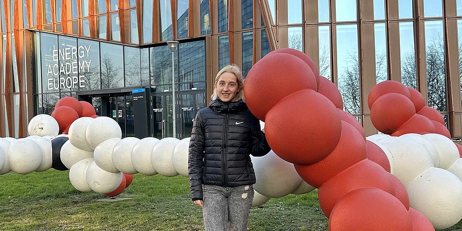 Eine Frau mit blonden Haaren lehnt sich an eine Modell eines Molekulars. Das Modell ist rot-weiß. 