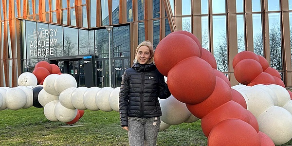Eine Frau mit blonden Haaren lehnt sich an eine Modell eines Molekulars. Das Modell ist rot-weiß. 