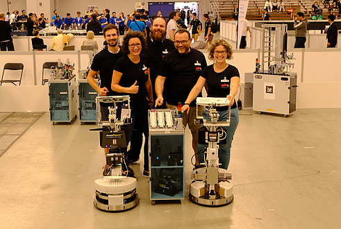 Three men and two women in black TU Graz T-shirts pose together with two robots in a production hall
