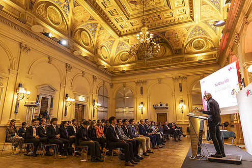 A man is standing at a lectern in a festive venue and speaking to an audience.