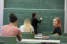 Three people are sitting in the rows of seats in a lecture theatre, while one person is writing something on the blackboard with chalk.
