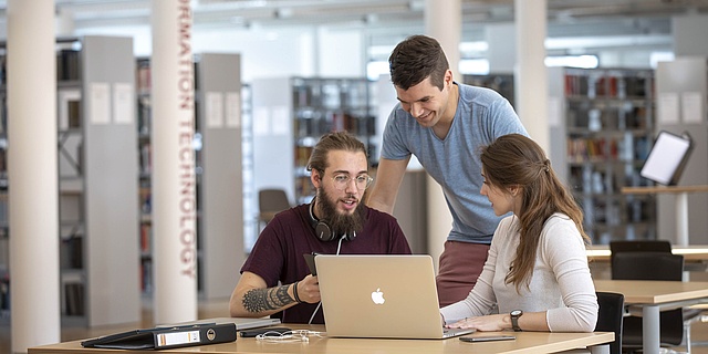 Zwei Studierende sitzen an einem Tisch. Eine Person steht hinter ihnen.