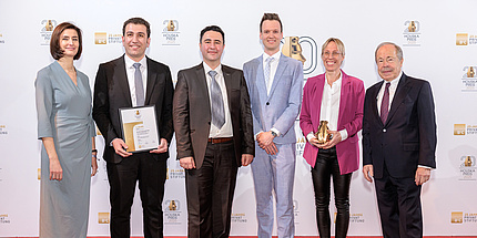 Six people smile at the camera. They are festively dressed. The second person on the right is holding a small trophy, the second person on the left a certificate.